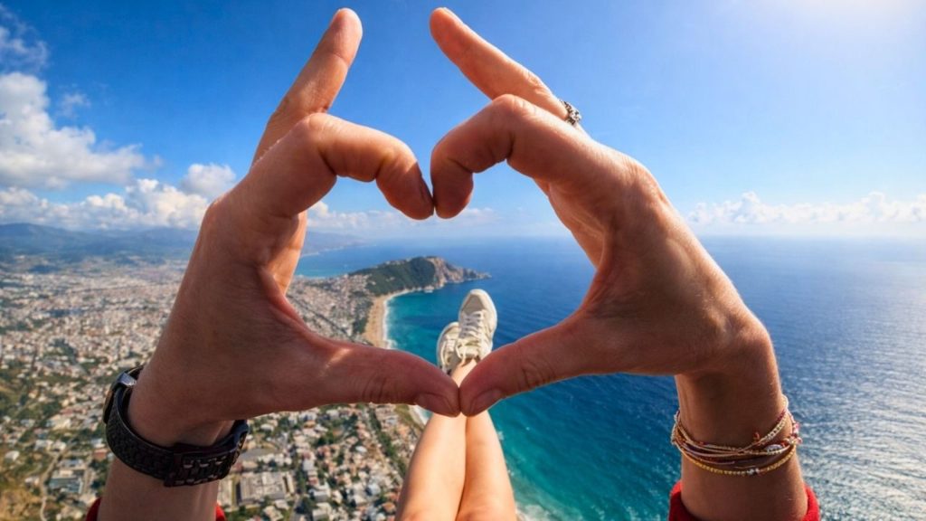 Heart shape hands during paragliding flight with Alanya Castle and Cleopatra Beach below