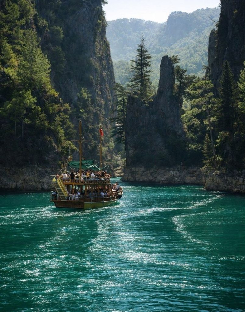 Wooden tour boat sailing between tall canyon walls on sparkling green water Oymapinar