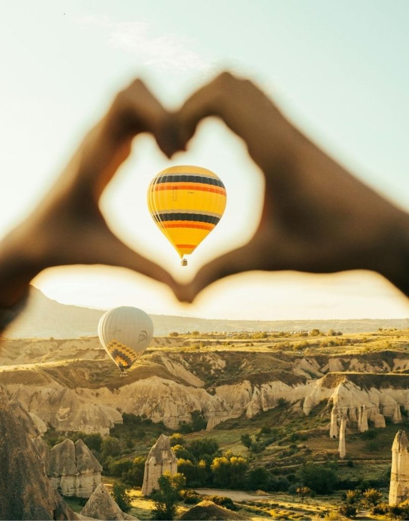 Heart-shaped hands framing hot air balloon over Cappadocia landscape at sunrise