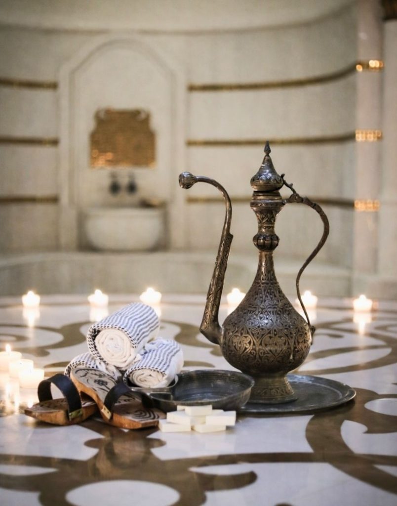 Traditional hammam accessories with Ottoman brass ibrik, candles, wooden clogs, and pestamal on marble floor
